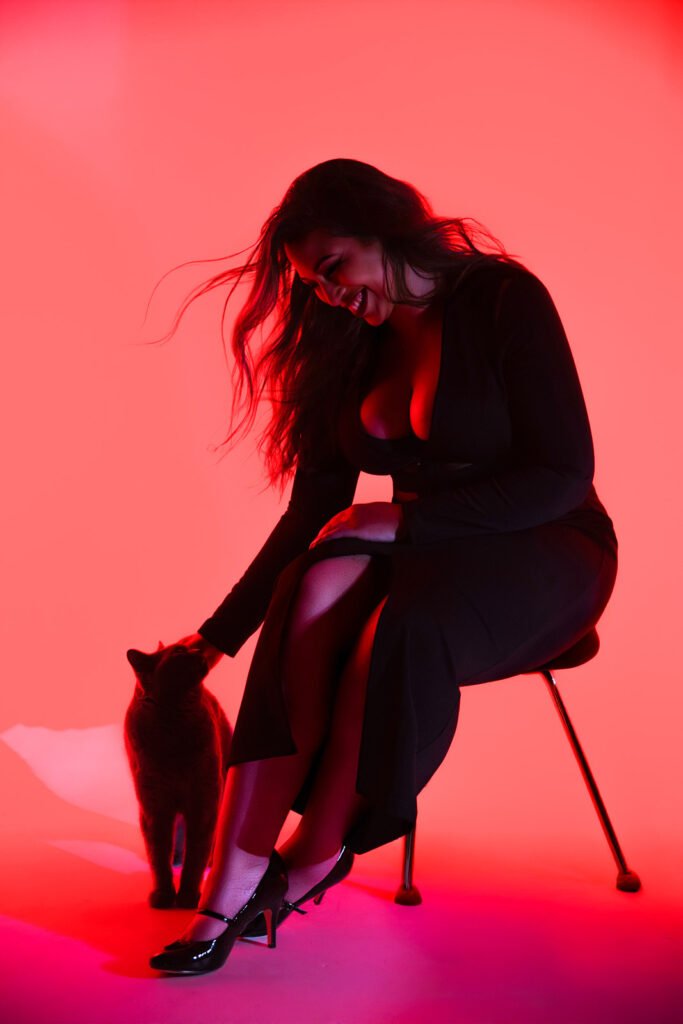 Seated portrait of laughing woman, pink lighting, black cat at her feet